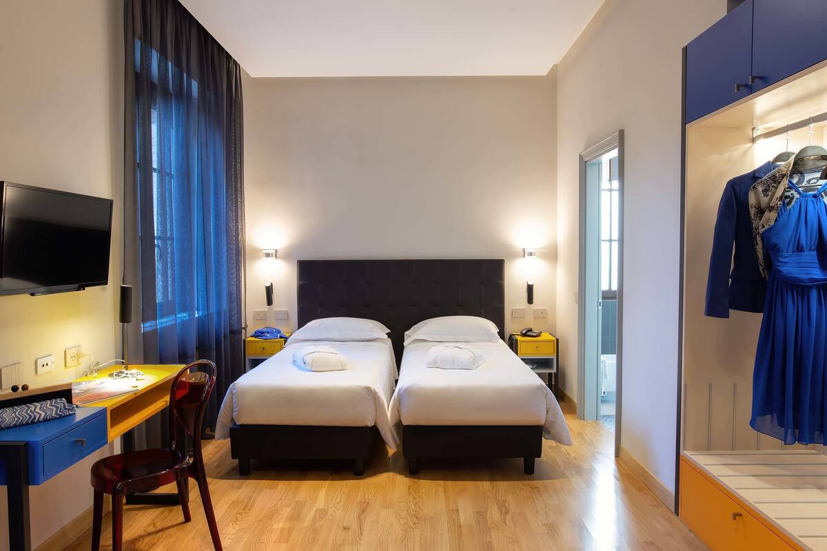 A hotel room with two beds, a desk, a TV, and a wardrobe with clothes.