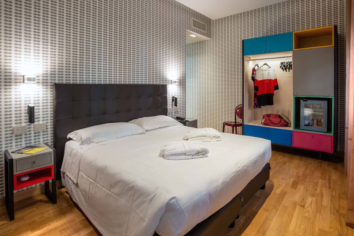 A hotel room with a large bed, a nightstand, a wardrobe, and a small seating area.