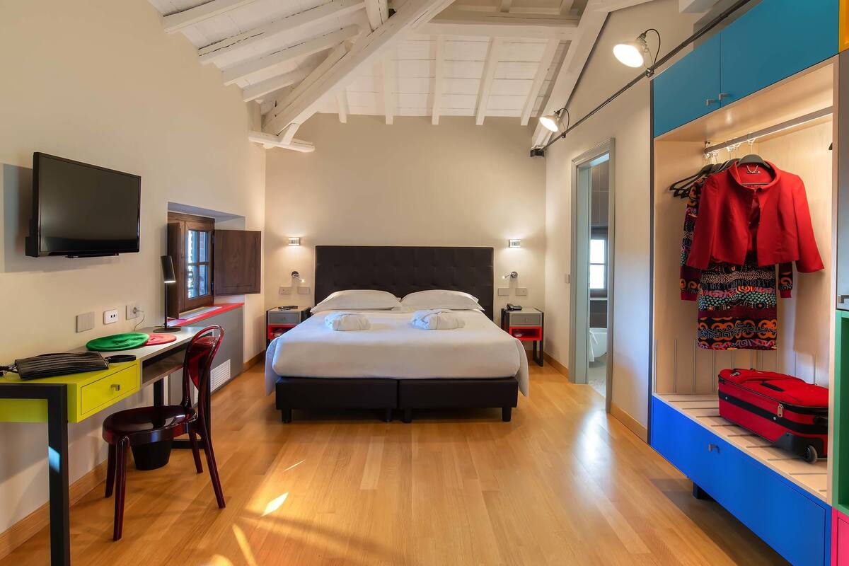A modern hotel room with a bed, desk, chair, wardrobe, and a TV.