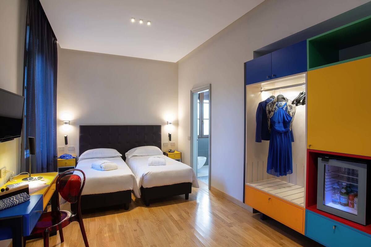 A hotel room with two beds, a desk with a chair, a wardrobe with a mirror, and a small refrigerator.