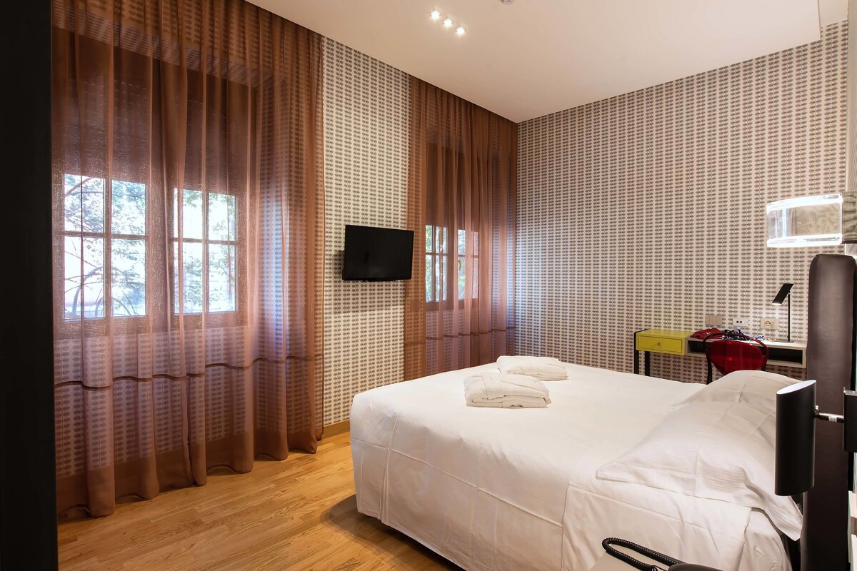 A hotel room with a large bed, a flat-screen TV, a desk with a chair, and a window with curtains.