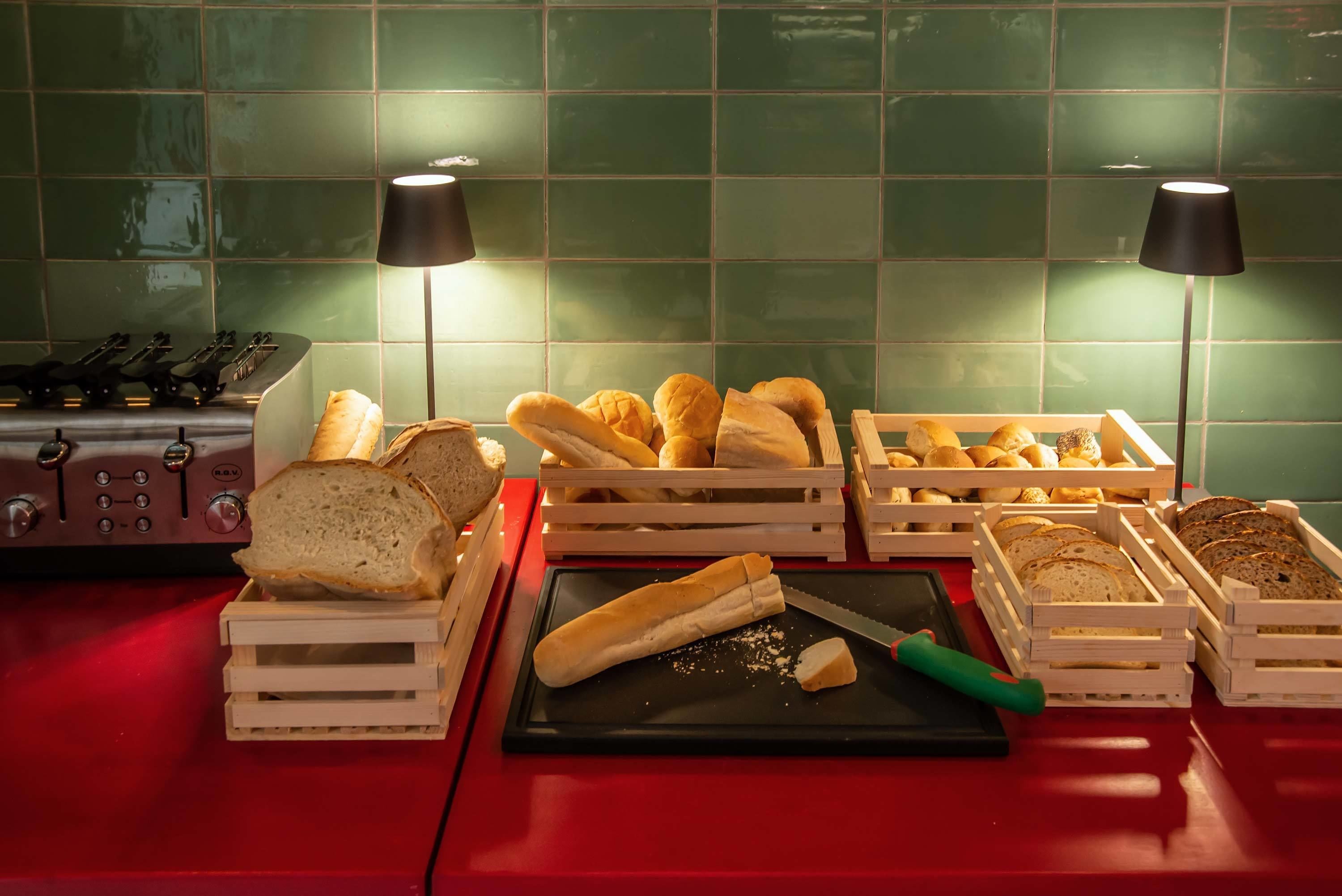 A kitchen counter with various bread types in wooden crates, a cutting board with a knife, and two lamps.