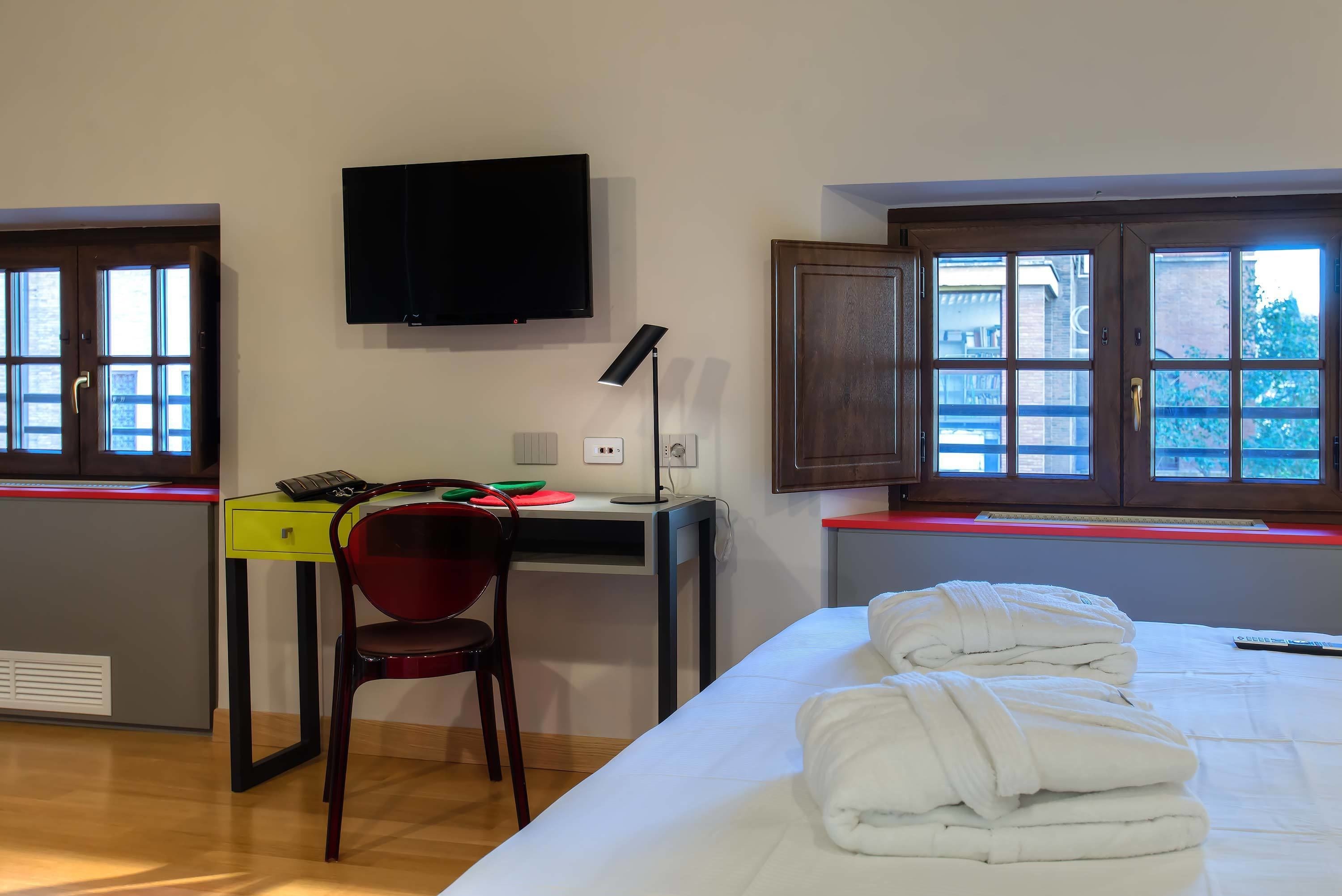 A hotel room with a bed, a desk with a chair, a television, and windows with blinds.
