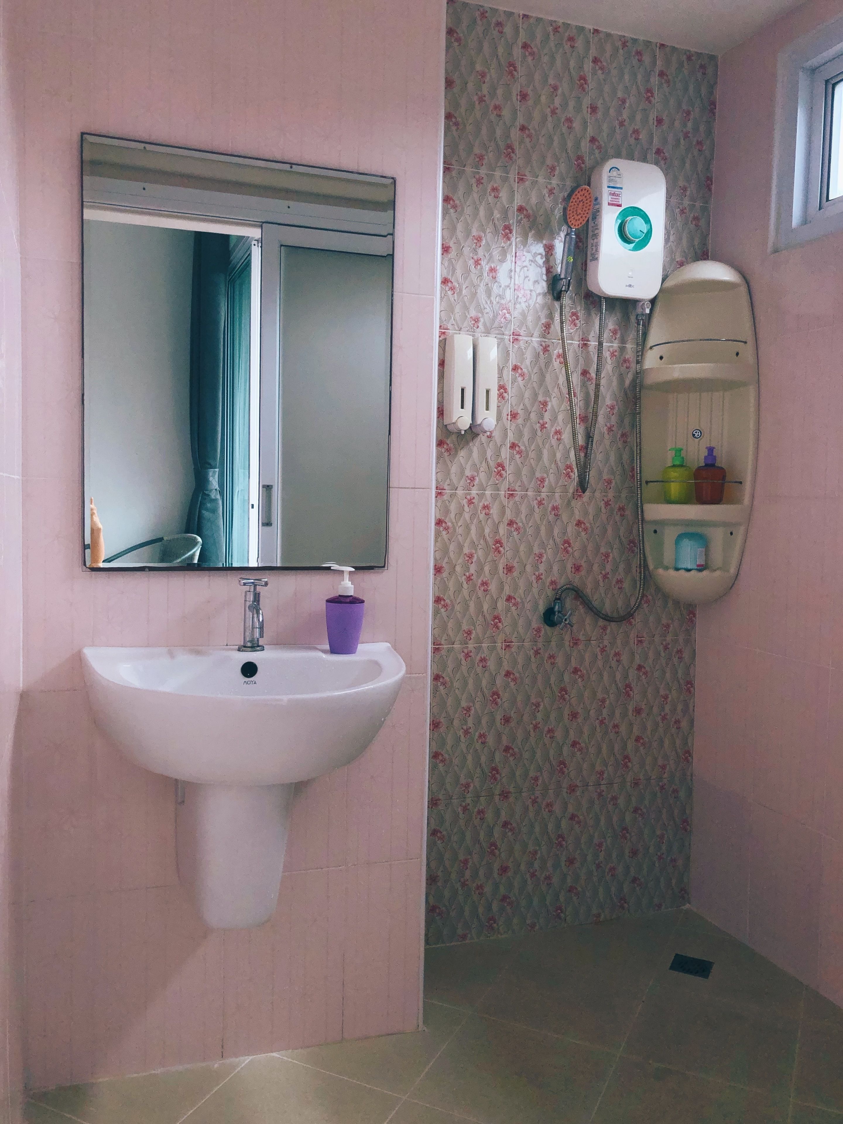 Un baño con un lavamanos blanco, un espejo, una ducha con detalles florales en la pared y un estante con artículos de aseo.