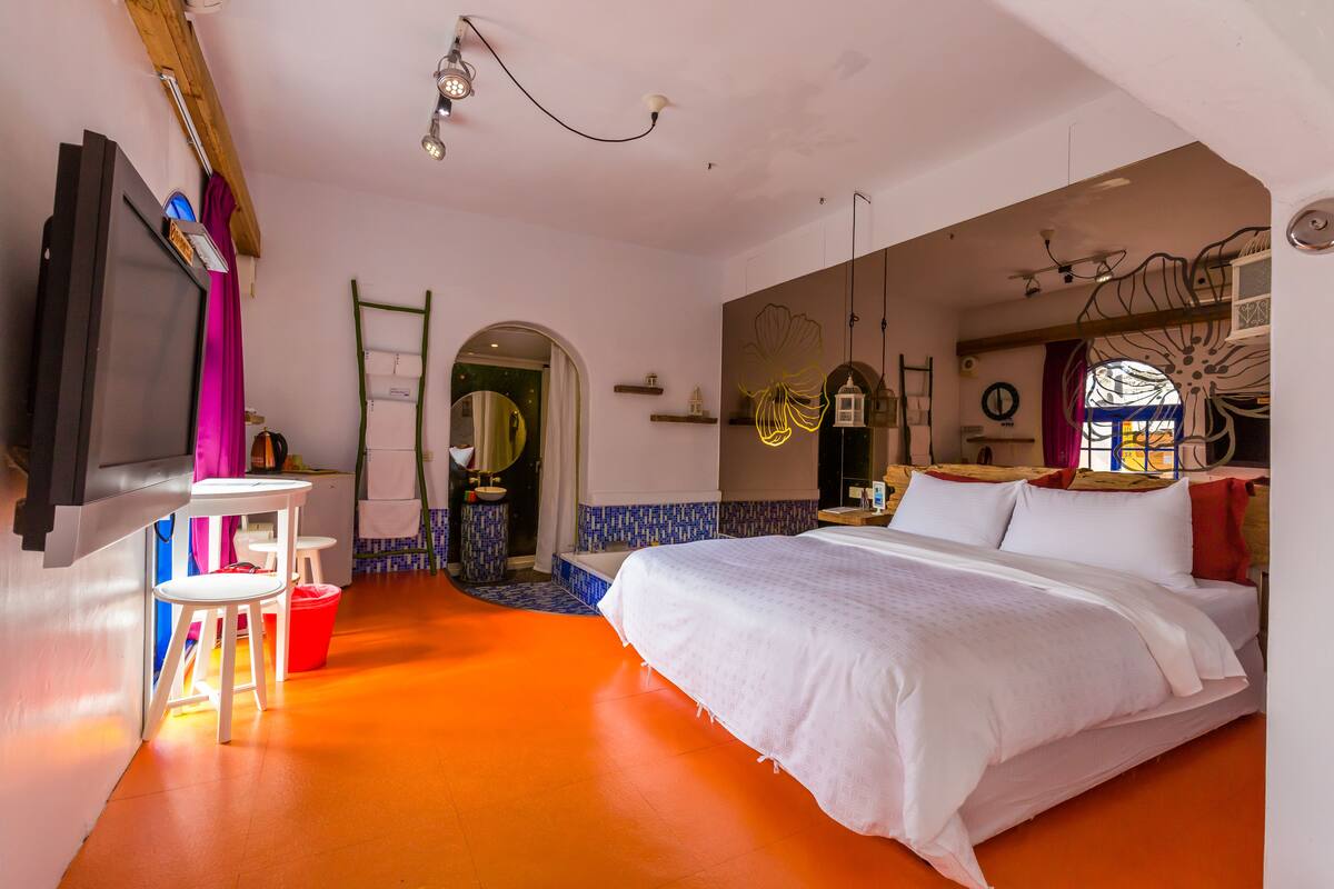 Un dormitorio con una cama grande, un televisor montado en la pared, una mesita con sillas y un baño visible a través de un arco.