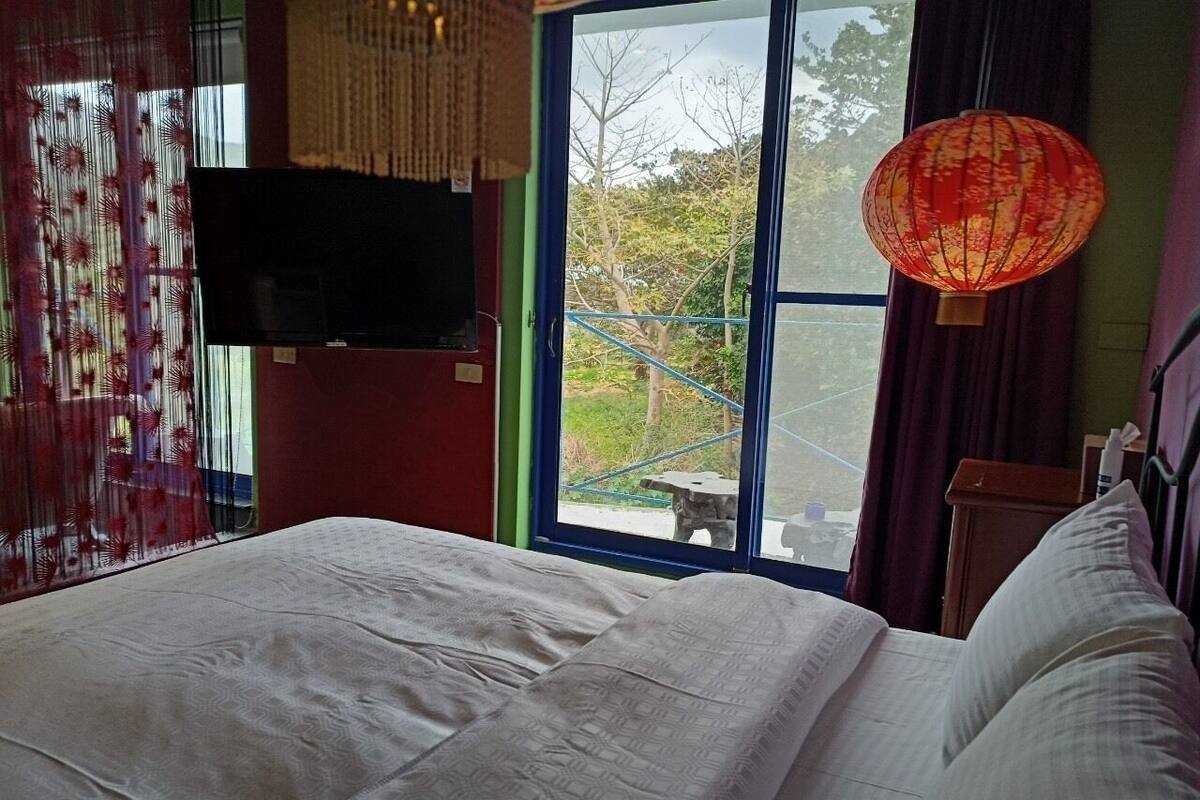Habitación de hotel con cama, televisión, ventana con vista a follaje y un farol rojo.