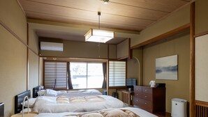 Ein traditionelles japanisches Schlafzimmer mit Tatami-Bodenbelag, einem großen Bett mit vielen Kissen, einem Schreibtisch mit Computer und Blick ins Freie durch Shoji-Schirme.