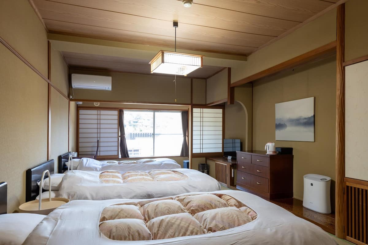 A traditional Japanese-style bedroom with tatami flooring, a large bed with multiple pillows, a desk with a computer, and a view of the outside through shoji screens.