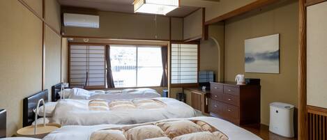 A traditional Japanese-style bedroom with tatami flooring, a large bed with multiple pillows, a desk with a computer, and a view of the outside through shoji screens.