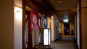 Ein Flur mit traditioneller japanischer Architektur, darunter Holzbalken und Tatami-Böden. Schilder weisen auf einen Wellnessbereich und einen Damenbereich hin.