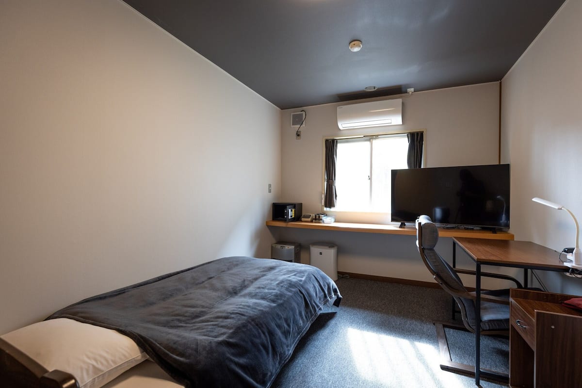 A compact hotel room with a bed, a desk with a computer, a TV, and a window with curtains.