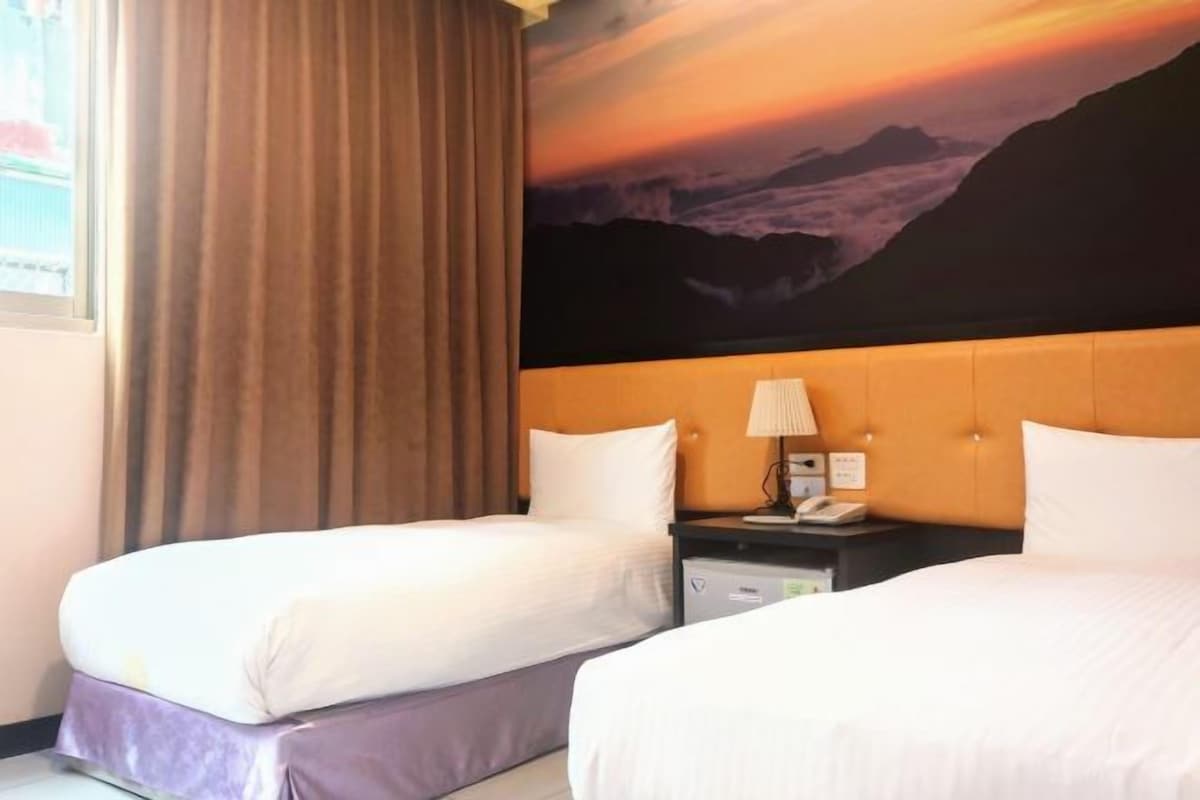 Une chambre d’hôtel avec deux lits, une table de chevet avec une lampe et un grand tableau représentant un paysage de montagne.