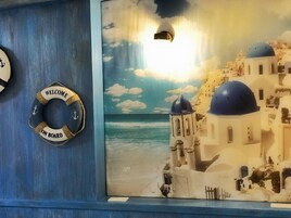 Una obra enmarcada de un pueblo costero con edificios blancos y cúpulas azules, un flotador con el texto 'WELCOME ON BOARD' y un pequeño bloque de madera sobre un estante azul.