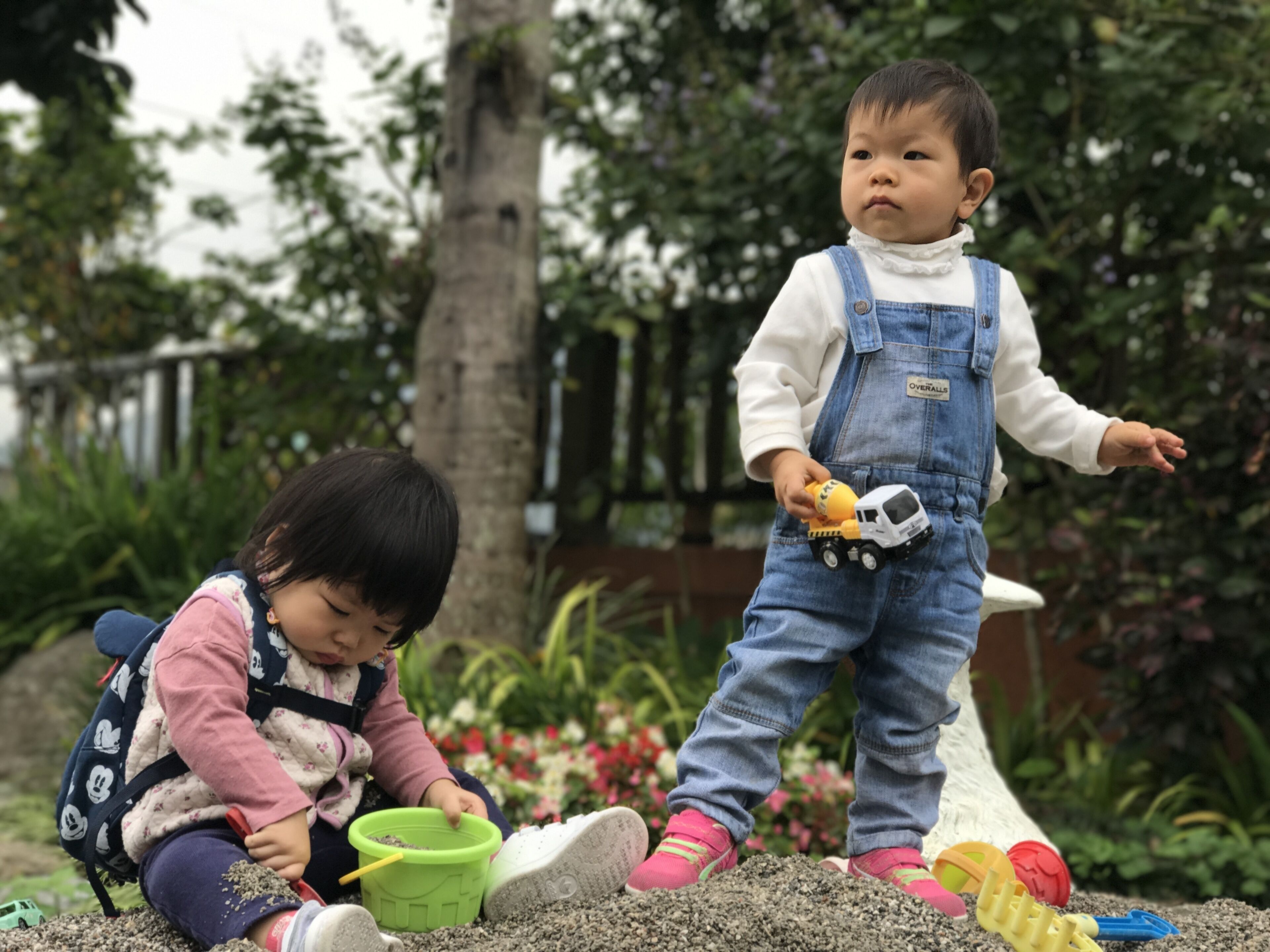 Two children playing with sand in an outdoor setting with greenery and a fence in the background.