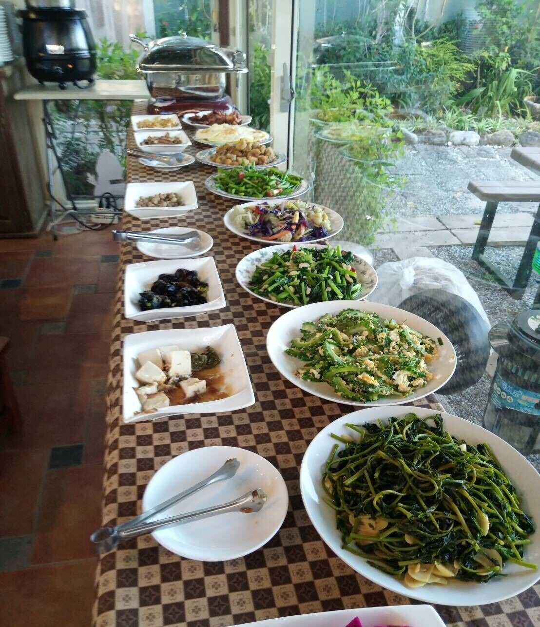 A buffet table with various dishes, including salads, meats, and vegetables, set up in a room with a checkered floor and a view of greenery outside.