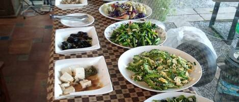 A buffet table with various dishes, including salads, meats, and vegetables, set up in a room with a checkered floor and a view of greenery outside.