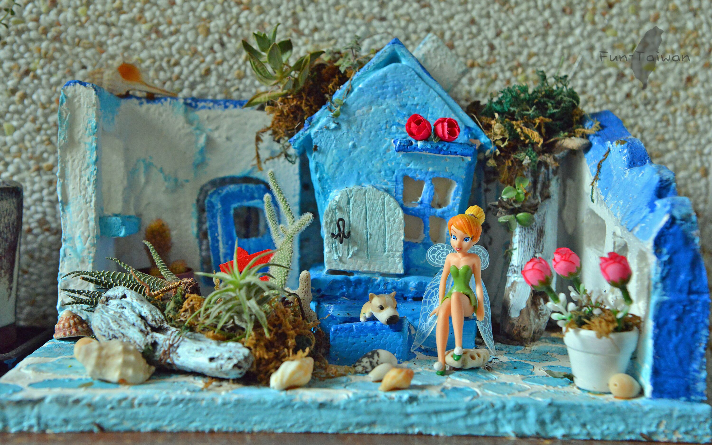 Una maqueta de una casa con una puerta azul, una figurita y diversas plantas.