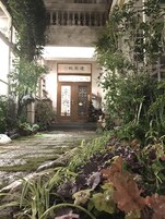Una entrada de edificio con puerta de madera, rodeada de abundante vegetación y plantas en macetas.