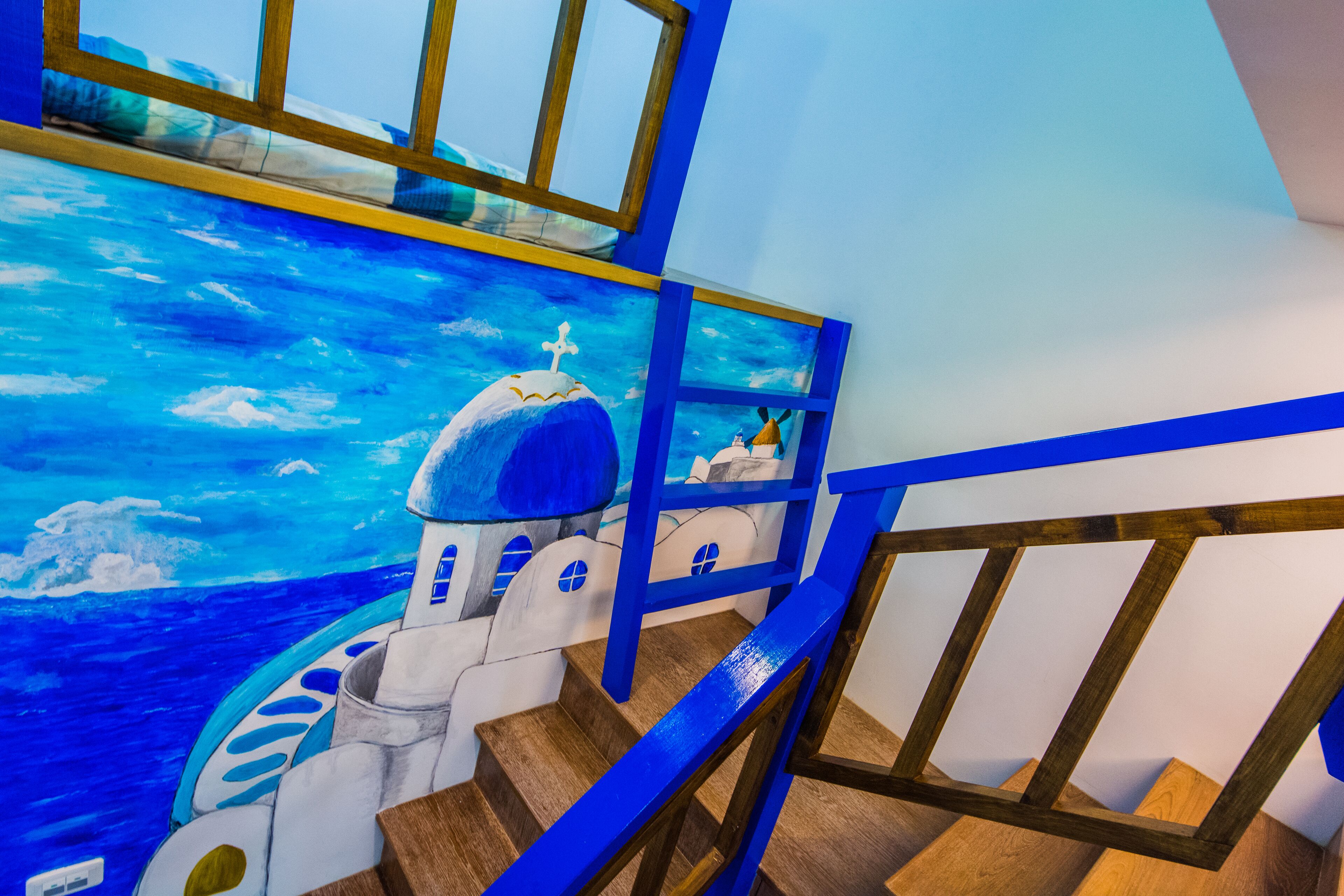 Escadaria com um mural de um edifício de cúpula azul e um navio, corrimãos de madeira e vista para o mar.