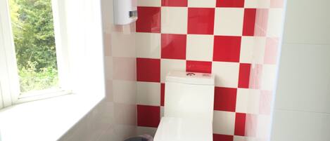 A bathroom with a red and white checkered wall pattern, a white toilet, a trash bin, and a window with a view of greenery.