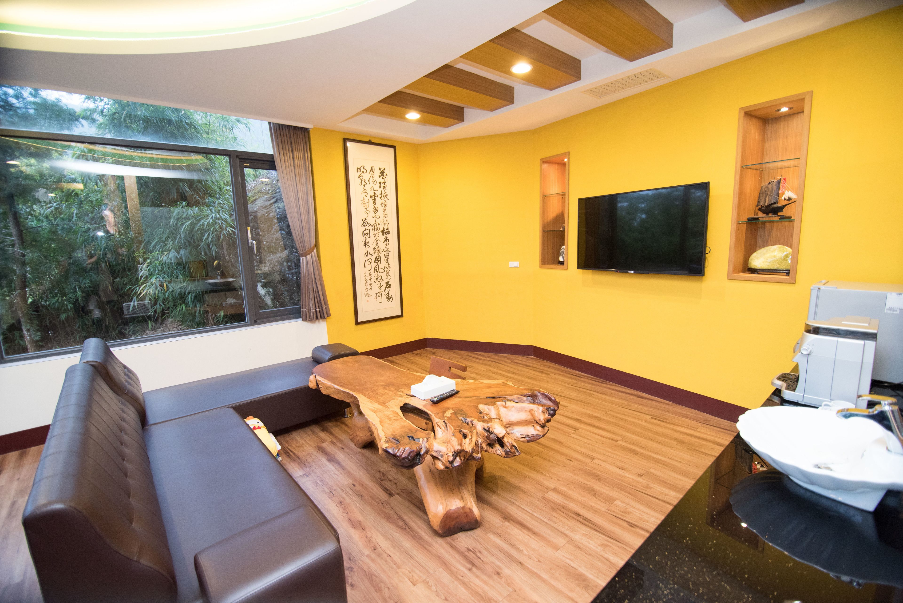 A modern living room with a large wooden coffee table, a sofa, a television mounted on the wall, and a window with a view of greenery.