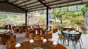 Un espace de restauration en plein air avec des tables rondes et des tabourets en bois, entouré de grandes fenêtres offrant une vue sur la verdure et un paysage lointain.