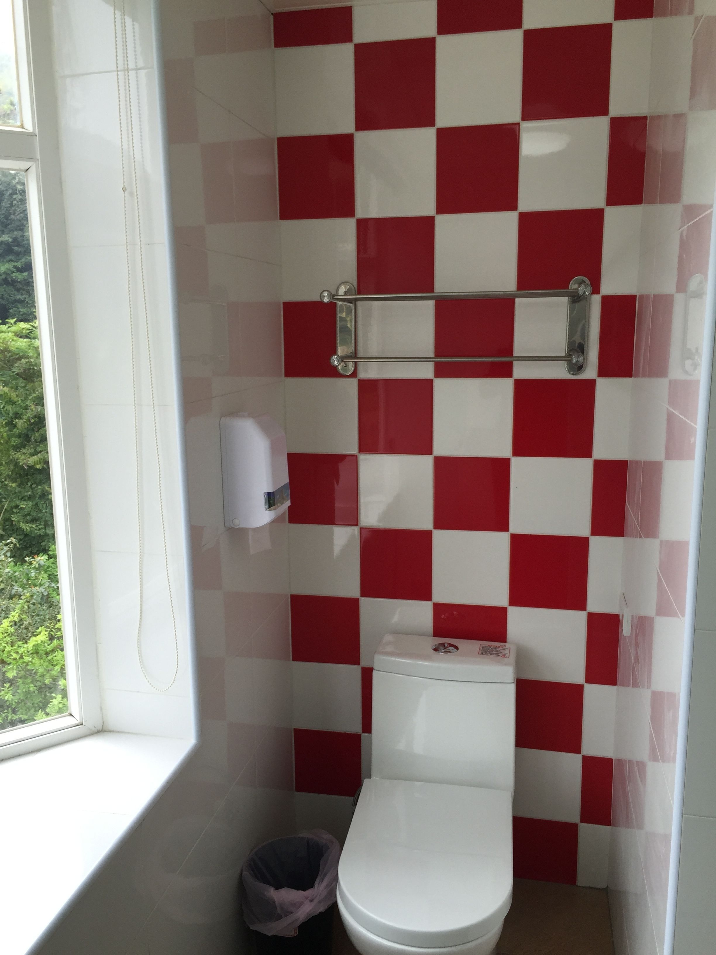 Une salle de bain avec un mur recouvert de carreaux rouges et blancs formant un motif à carreaux, des toilettes blanches, une poubelle et une fenêtre donnant sur un paysage verdoyant.
