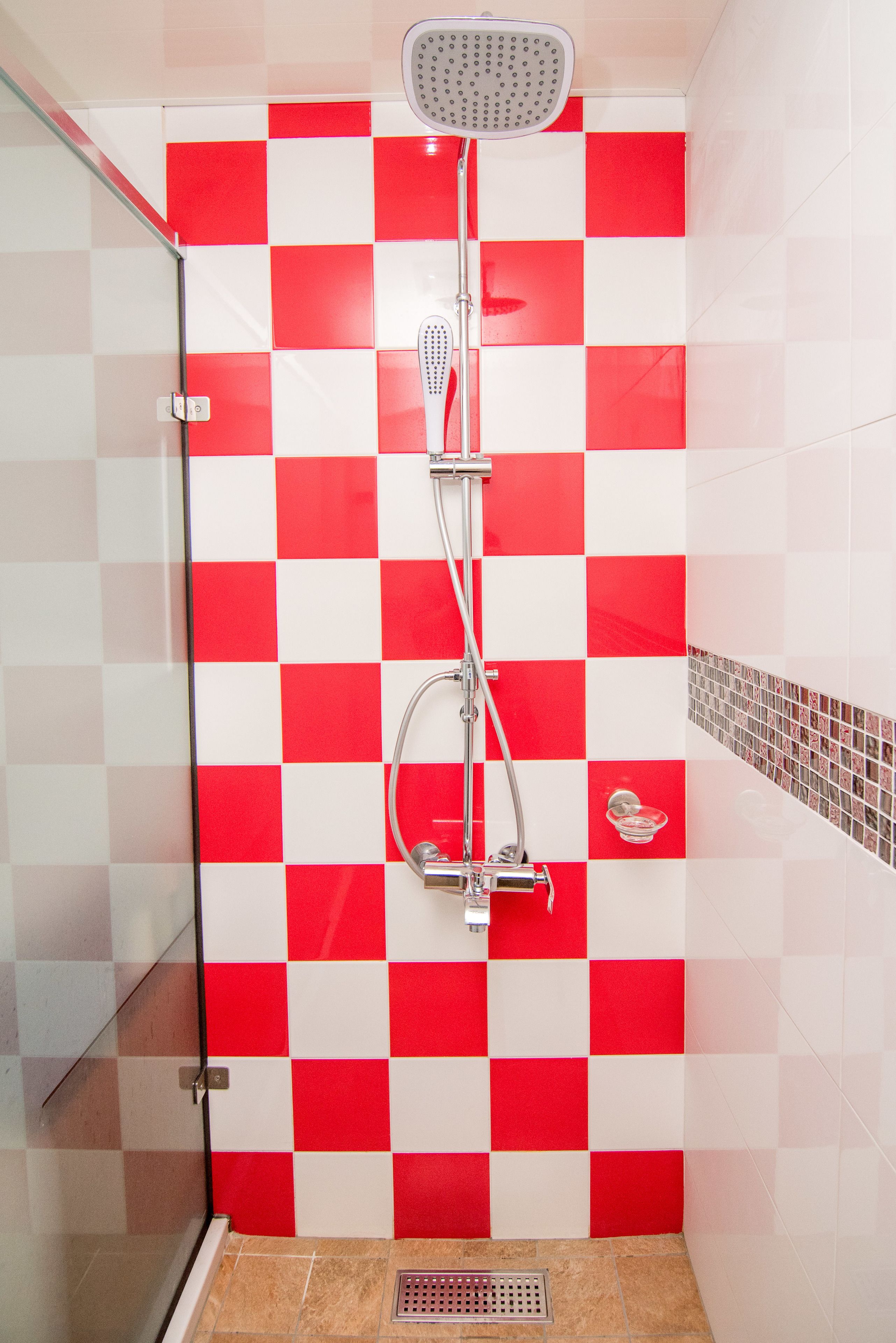 Une douche avec un mur à motifs de carreaux rouges et blancs, une porte en verre et un pommeau de douche.