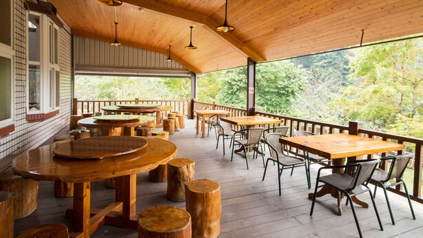 Une terrasse extérieure en bois, dotée de tables et de chaises, entourée d’une balustrade en bois et offrant une vue sur une zone boisée.