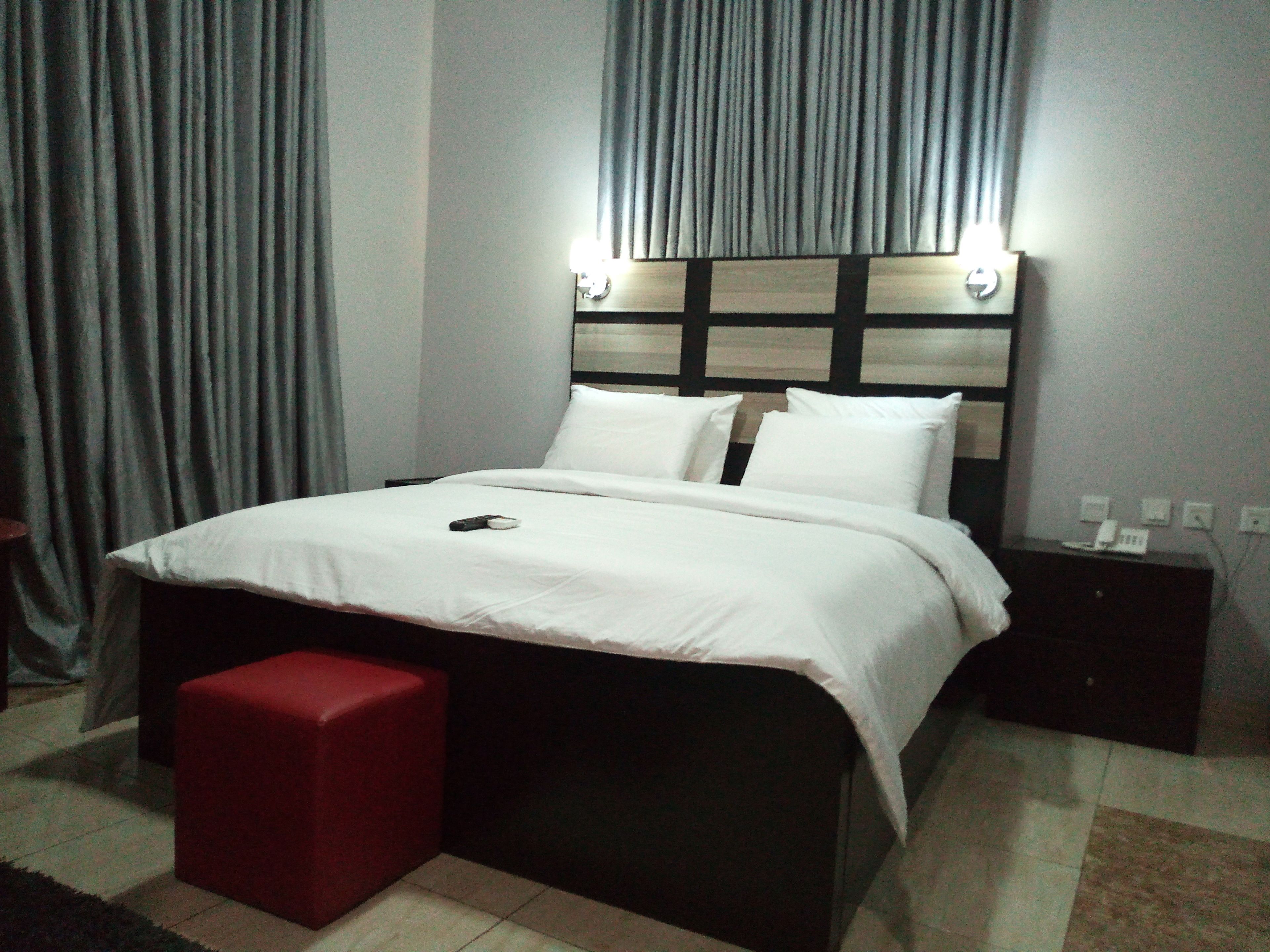 A neatly made bed with white linens, a red ottoman, and a bedside table with a lamp.