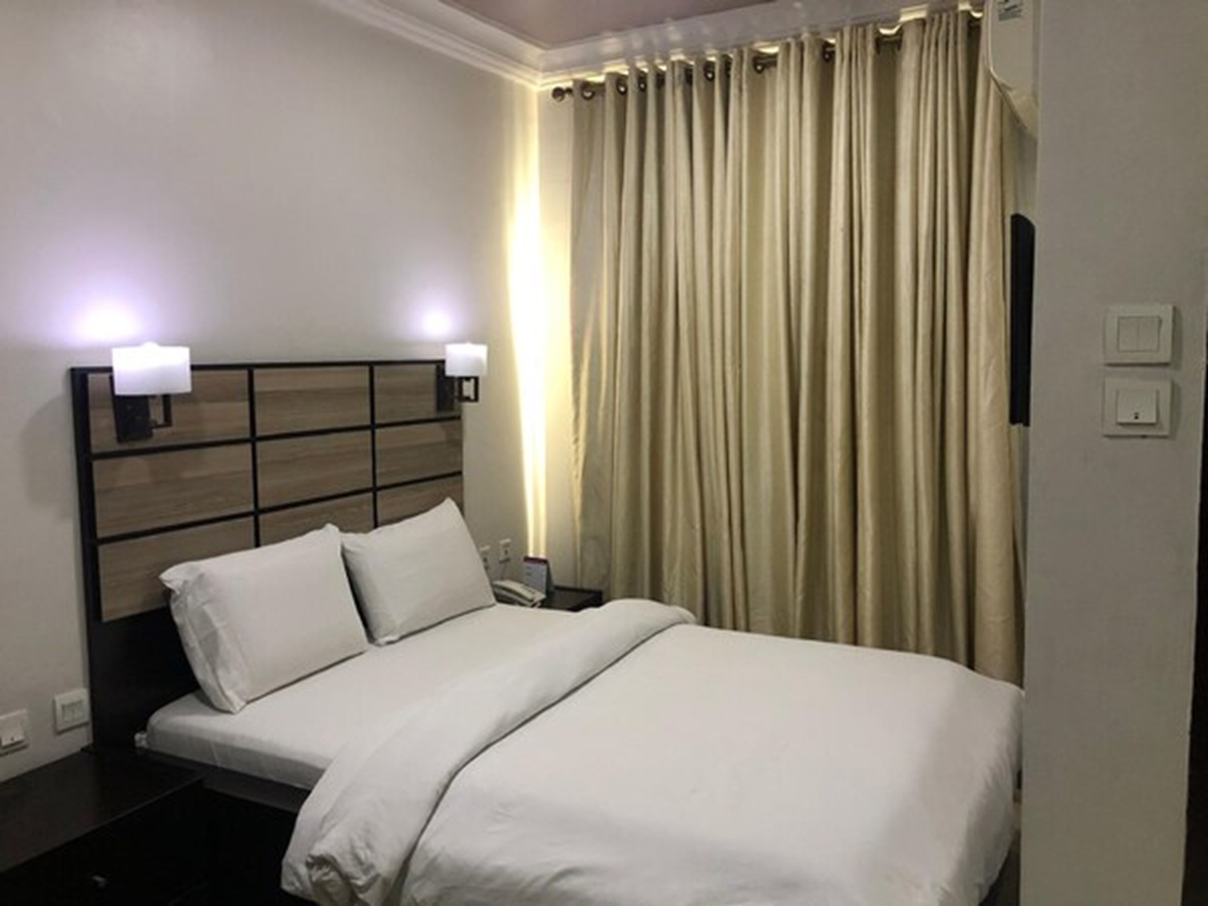 A hotel room with a bed, two bedside lamps, a nightstand, and a curtain.