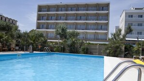 Una piscina con una escalera metálica, rodeada por un suelo de baldosas. Al fondo, un edificio de varios pisos con balcones.