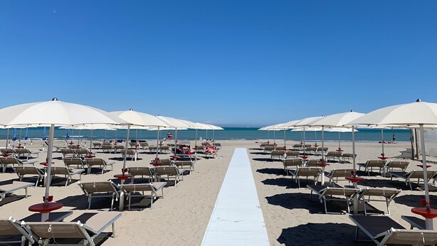 Un hotel frente a la playa con filas de tumbonas y sombrillas, un paseo marítimo blanco que conduce al mar, y un cielo azul despejado.