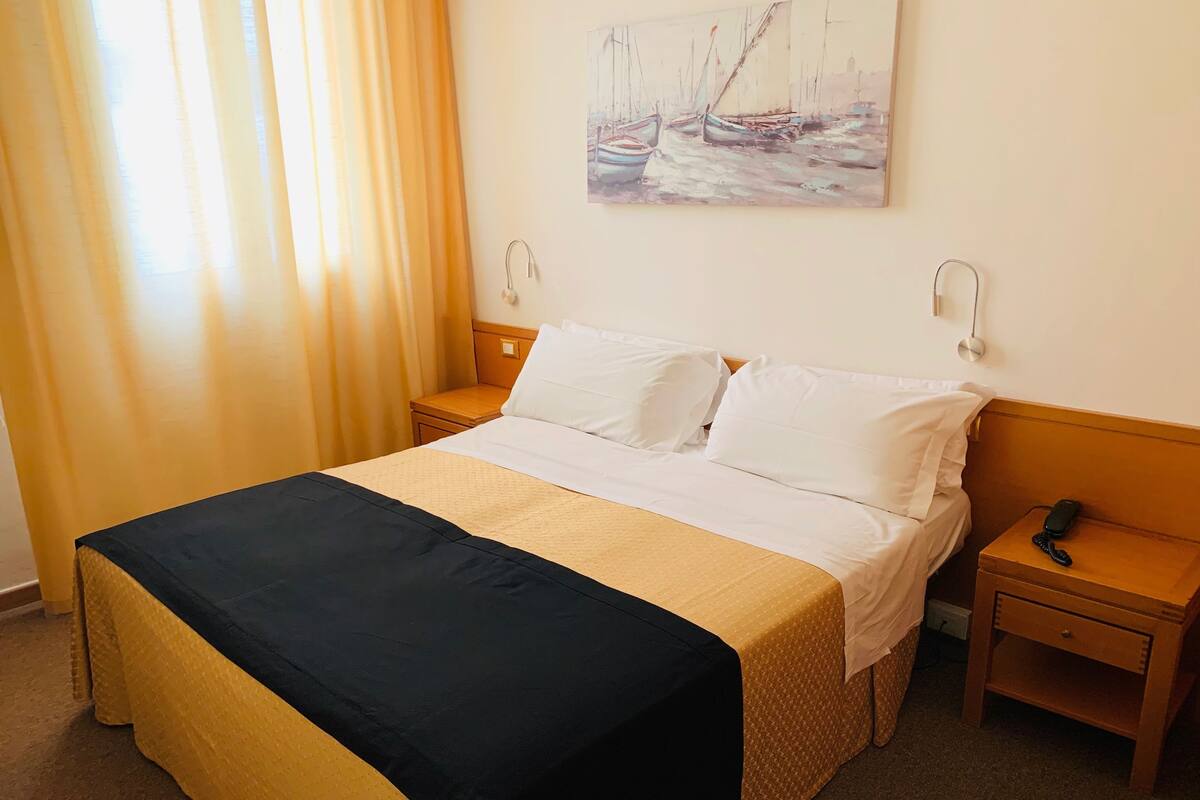 A hotel room with a bed, two bedside tables, a nightstand, a painting on the wall, and a window with curtains.