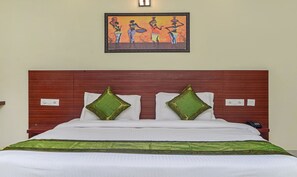 A hotel room with a wooden headboard, a bed with white bedding and green decorative pillows, a painting above the bed, and a bedside table with a phone.