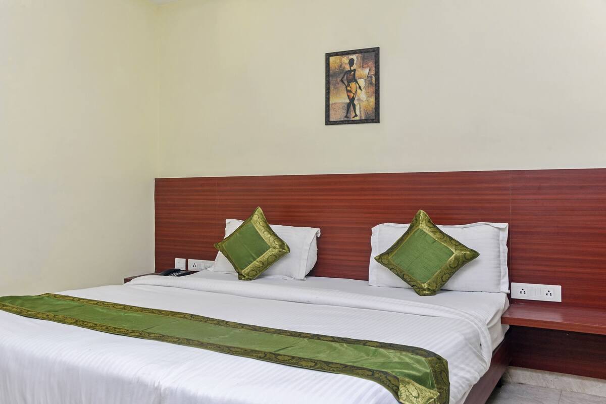 A hotel room with a wooden headboard, a bed with white bedding and green decorative pillows, a green throw blanket, and a painting on the wall.