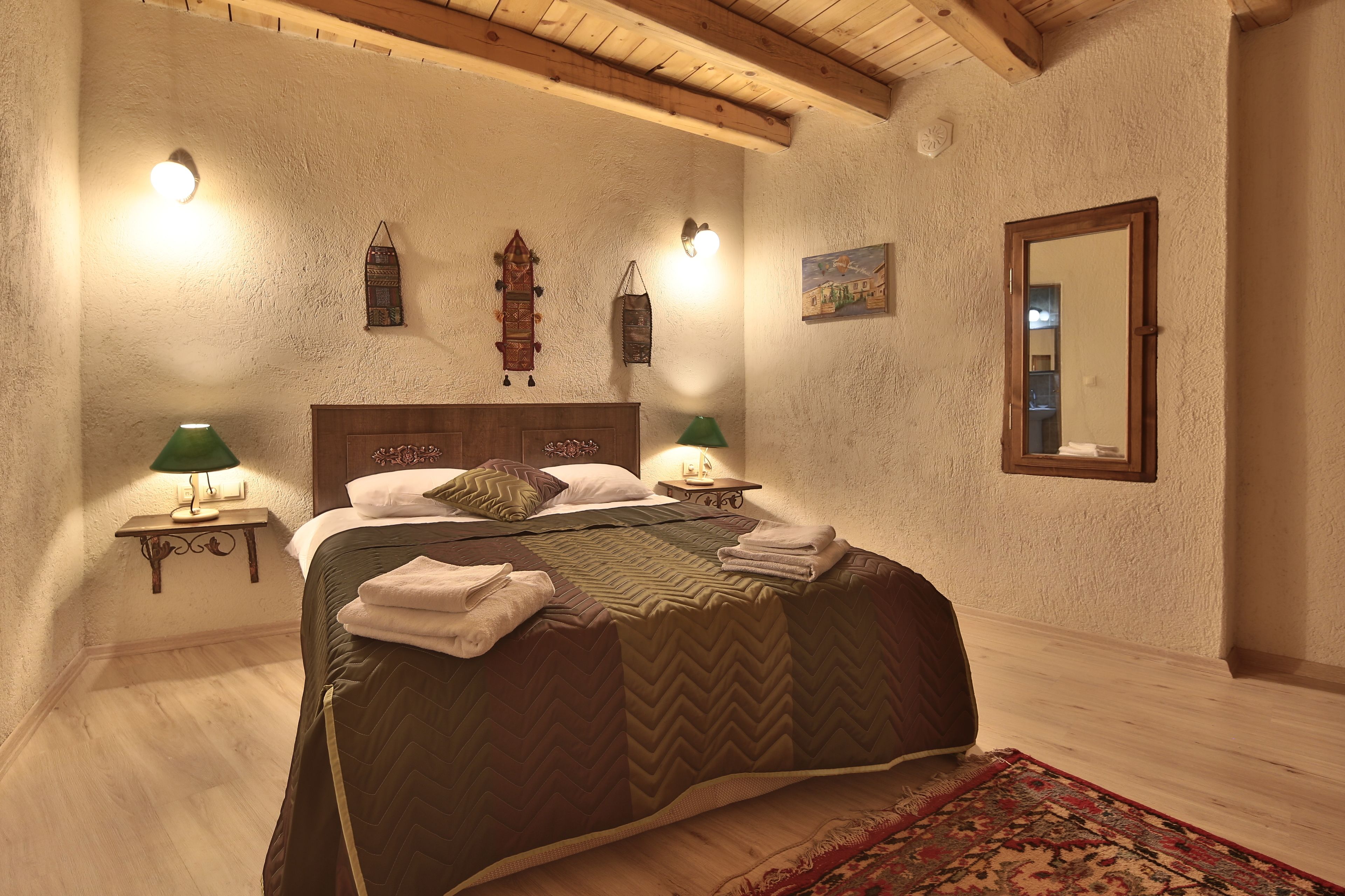 A bedroom with a wooden ceiling, a bed with a green quilt, two bedside lamps, a wall-mounted clock, and a mirror.