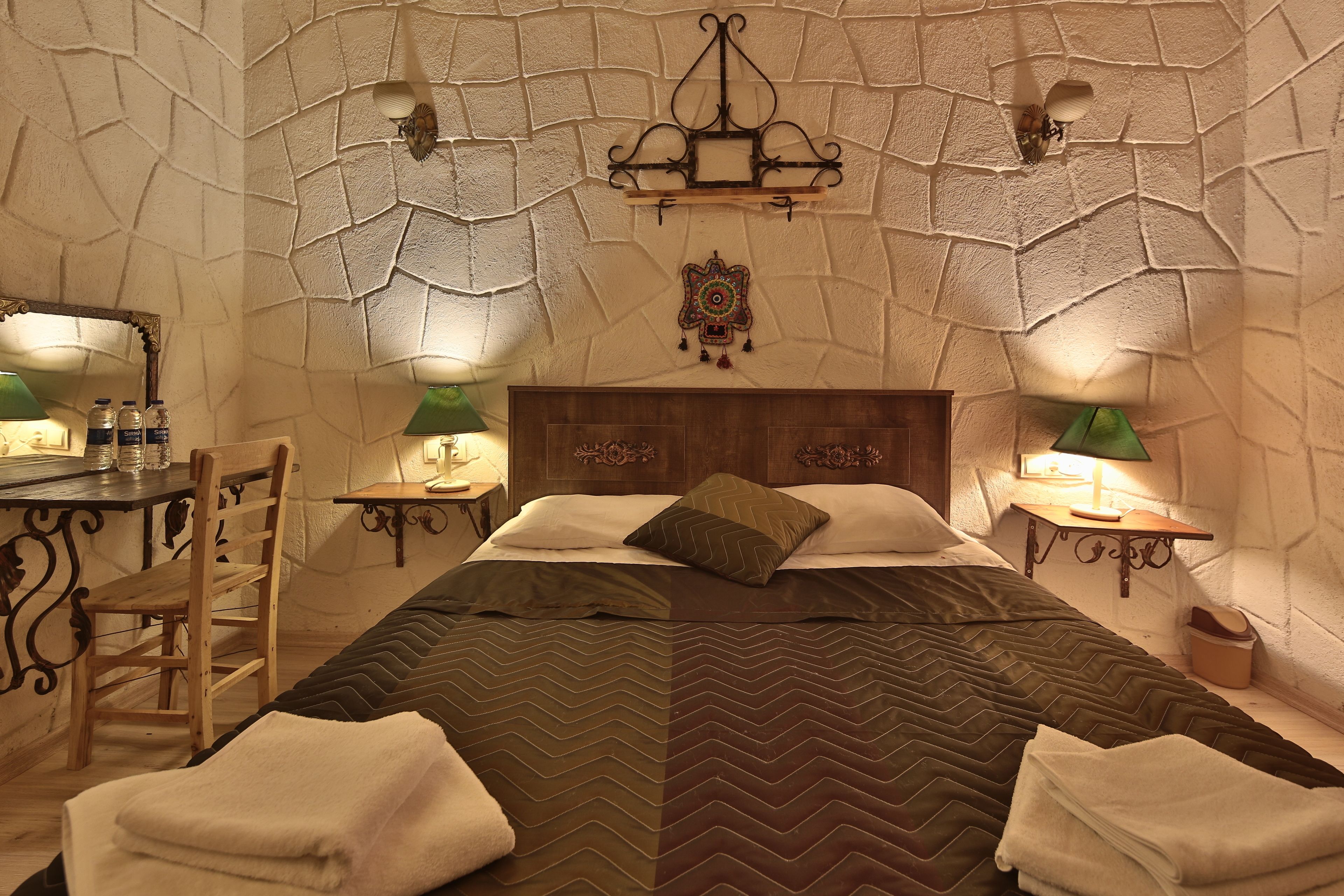 A bedroom with a stone wall, a wooden bed, two bedside tables with lamps, a mirror, and a chair.
