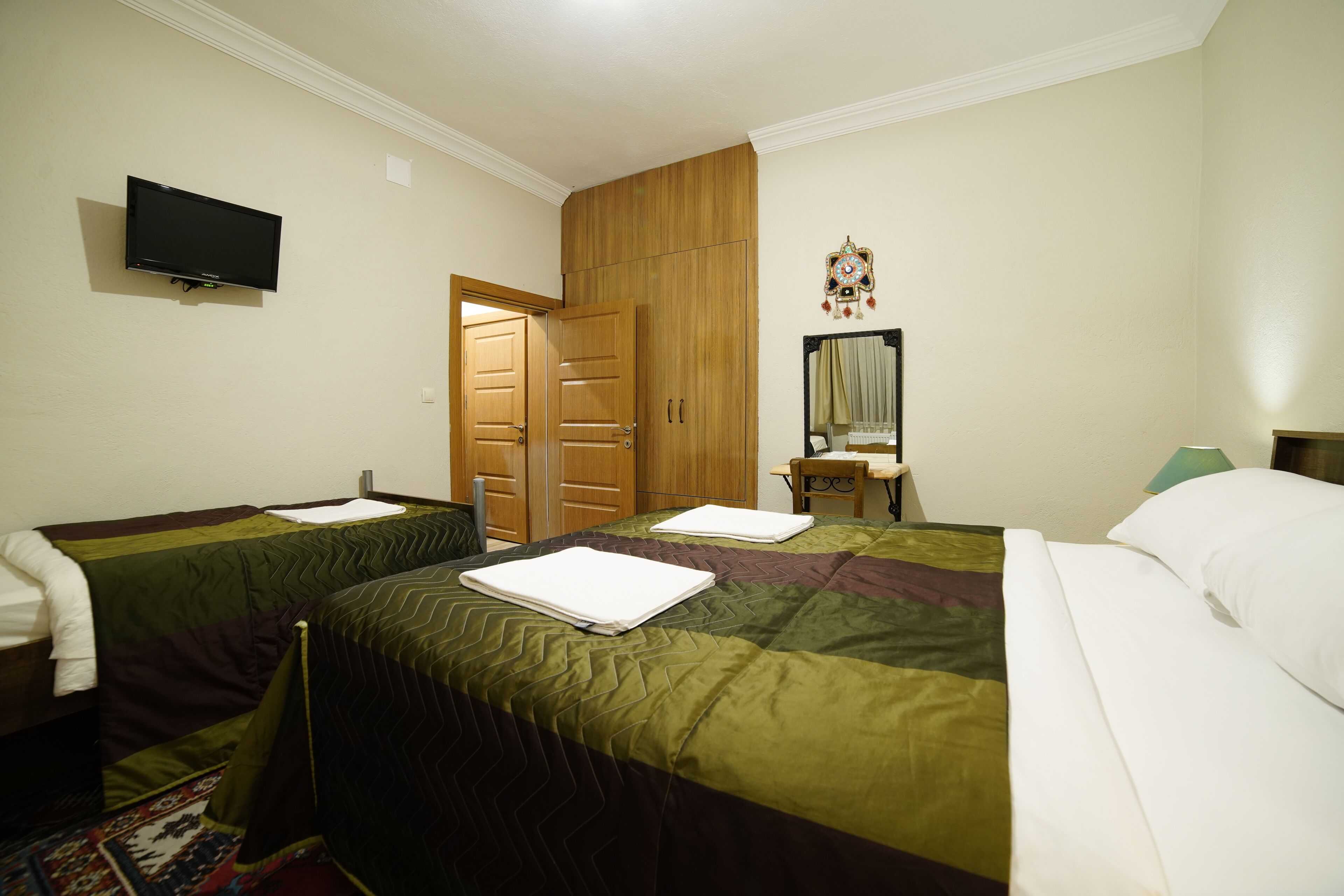 A hotel room with two beds, a television, a wooden wardrobe, a mirror, and a desk with a lamp.