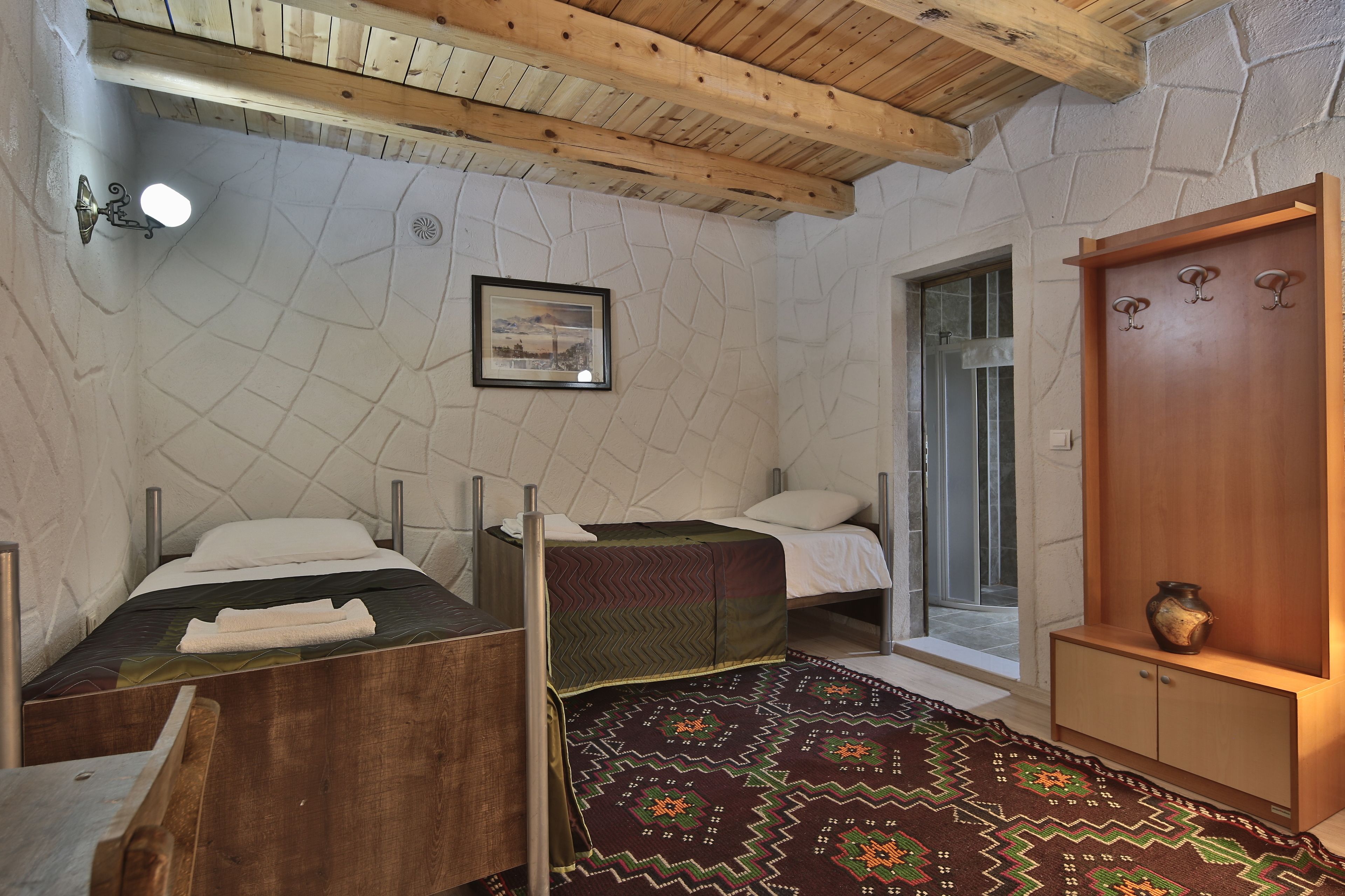 Una habitación con dos camas, techo de madera, pared de piedra, un cuadro enmarcado, un armario de madera y una alfombra estampada.