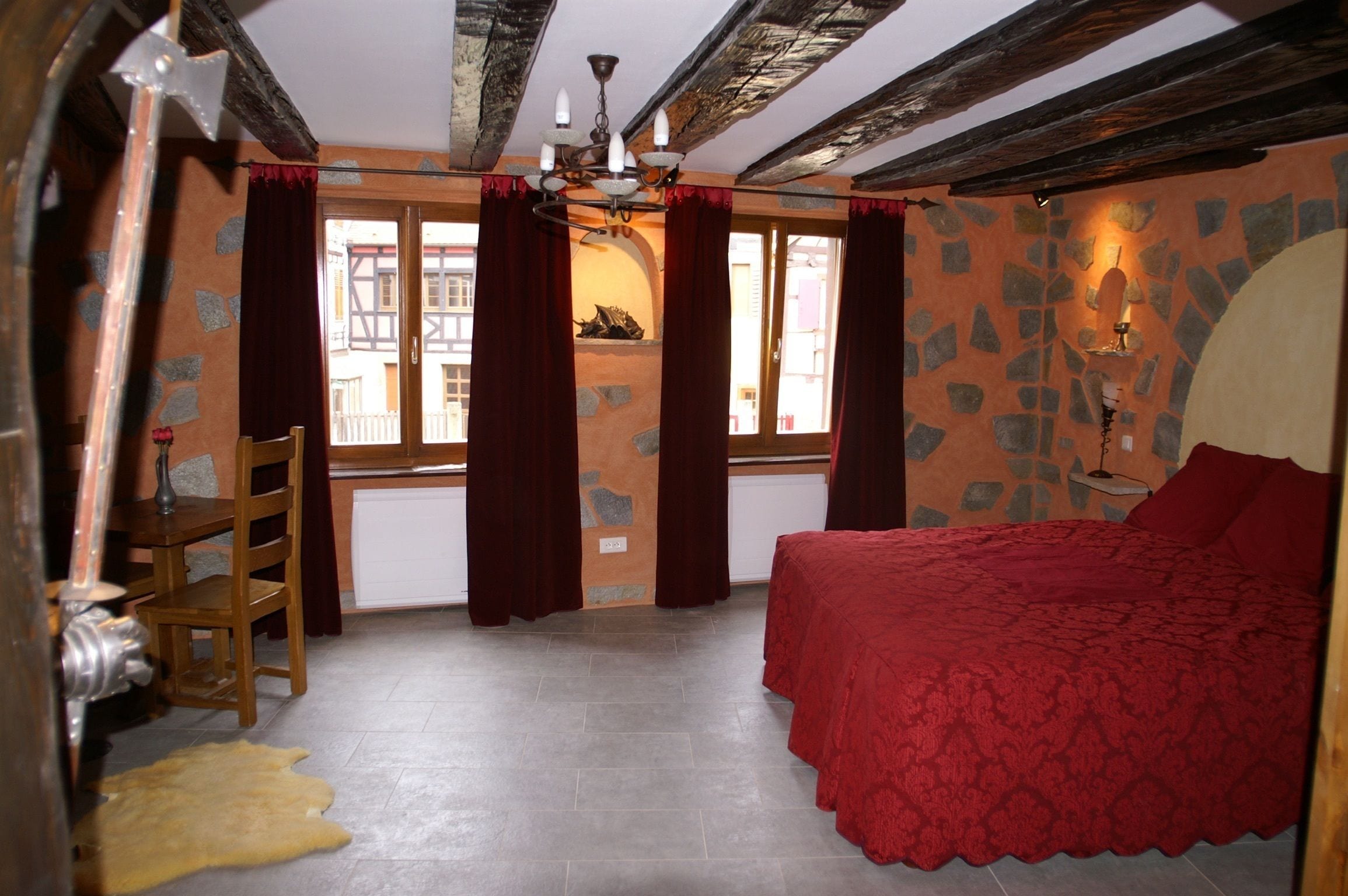 A bedroom with a bed, a chair, a window with curtains, a stone wall, and a chandelier.