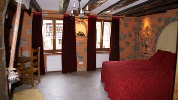 A bedroom with a bed, a chair, a window with curtains, a stone wall, and a chandelier.