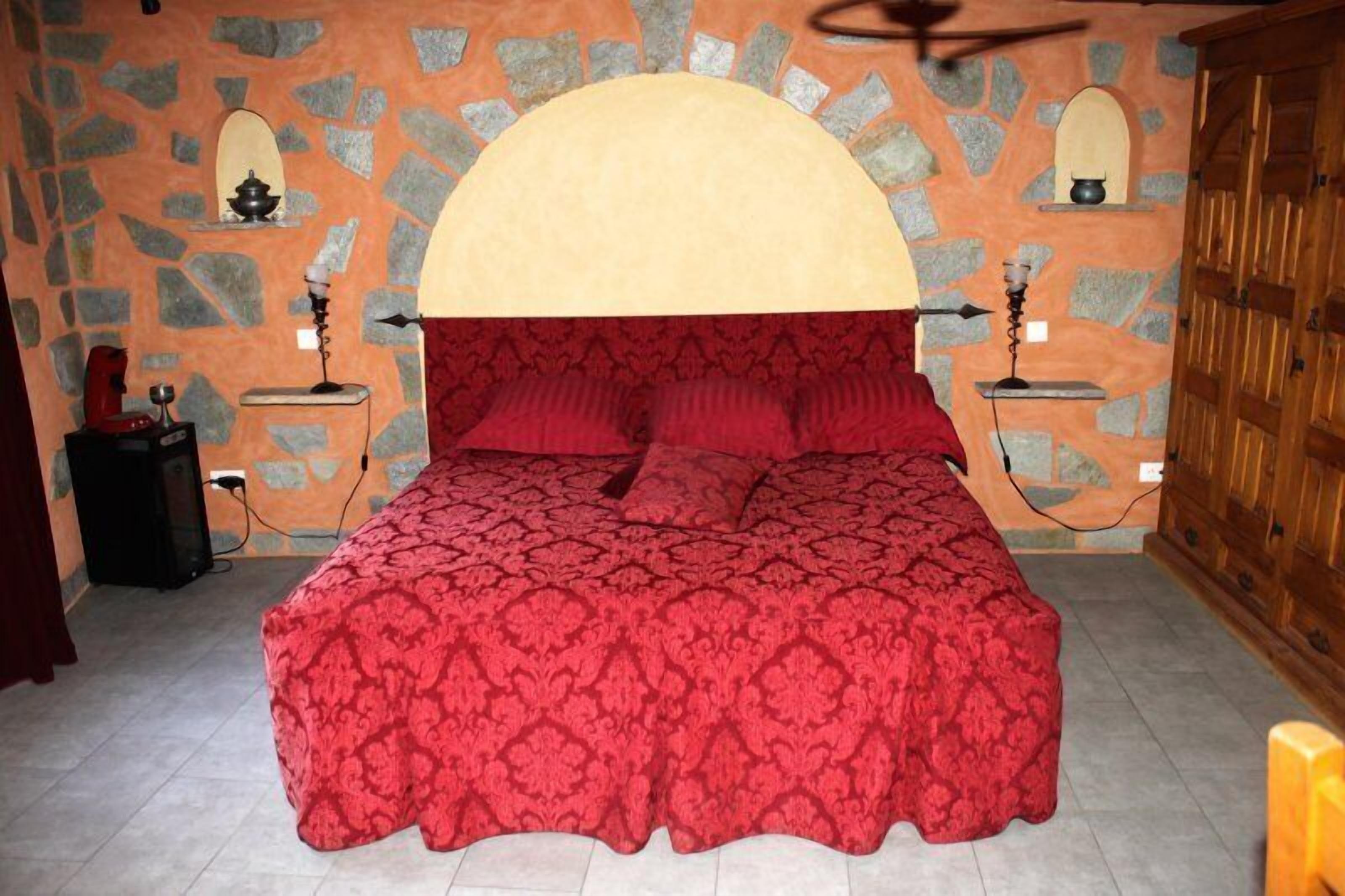 A bedroom with a stone wall, a bed with a red bedspread, two bedside tables with lamps, and a wooden wardrobe.