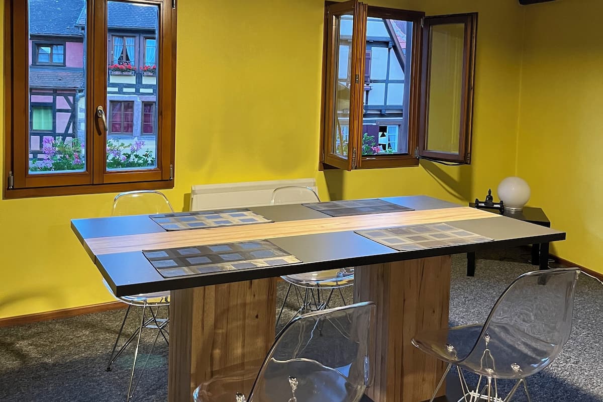 Una habitación con una mesa de madera y sillas transparentes, paredes amarillas y vigas a la vista en el techo.