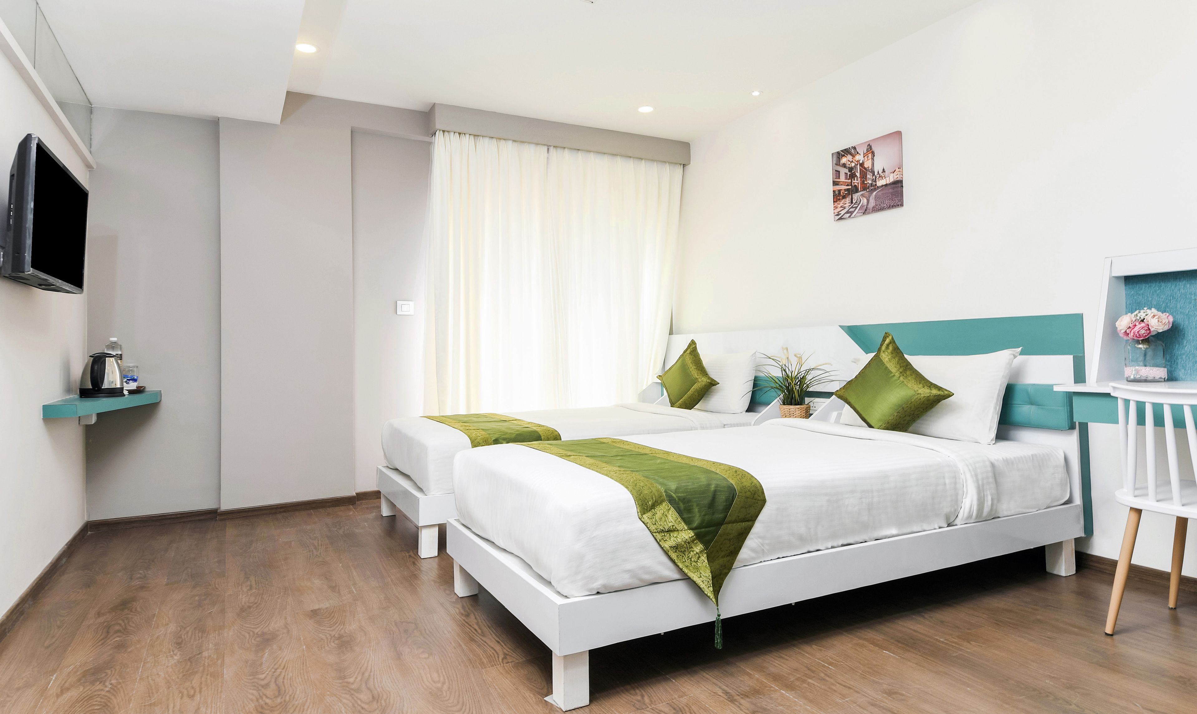 A modern hotel room with two single beds, a small desk, a flat-screen TV, and a window with curtains.
