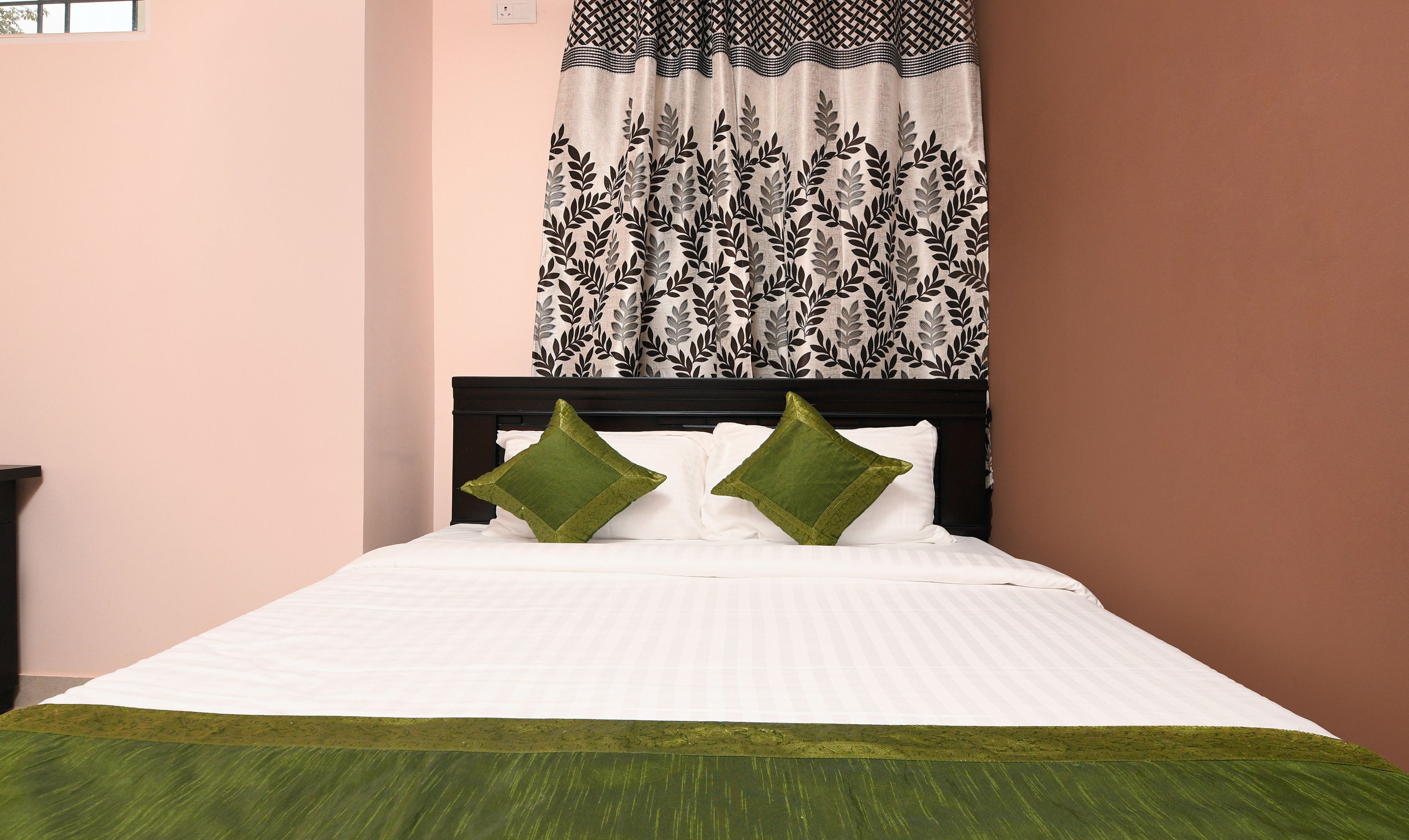 A bed with white bedding and two green pillows, a dark headboard, and a patterned curtain.