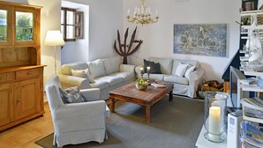 Una sala de estar acogedora con una mesa de centro de madera, dos sofás blancos, un sillón a cuadros, una araña y un cuadro abstracto grande en la pared.
