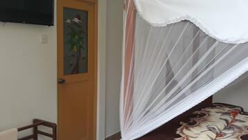 A bedroom with a bed under a white canopy, a wooden door with a floral painting, and a TV mounted on the wall.