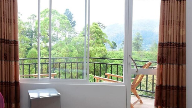 A room with a large window offering a view of a balcony with wooden furniture and a railing. - Lilly Guest Inn (Haputhale)