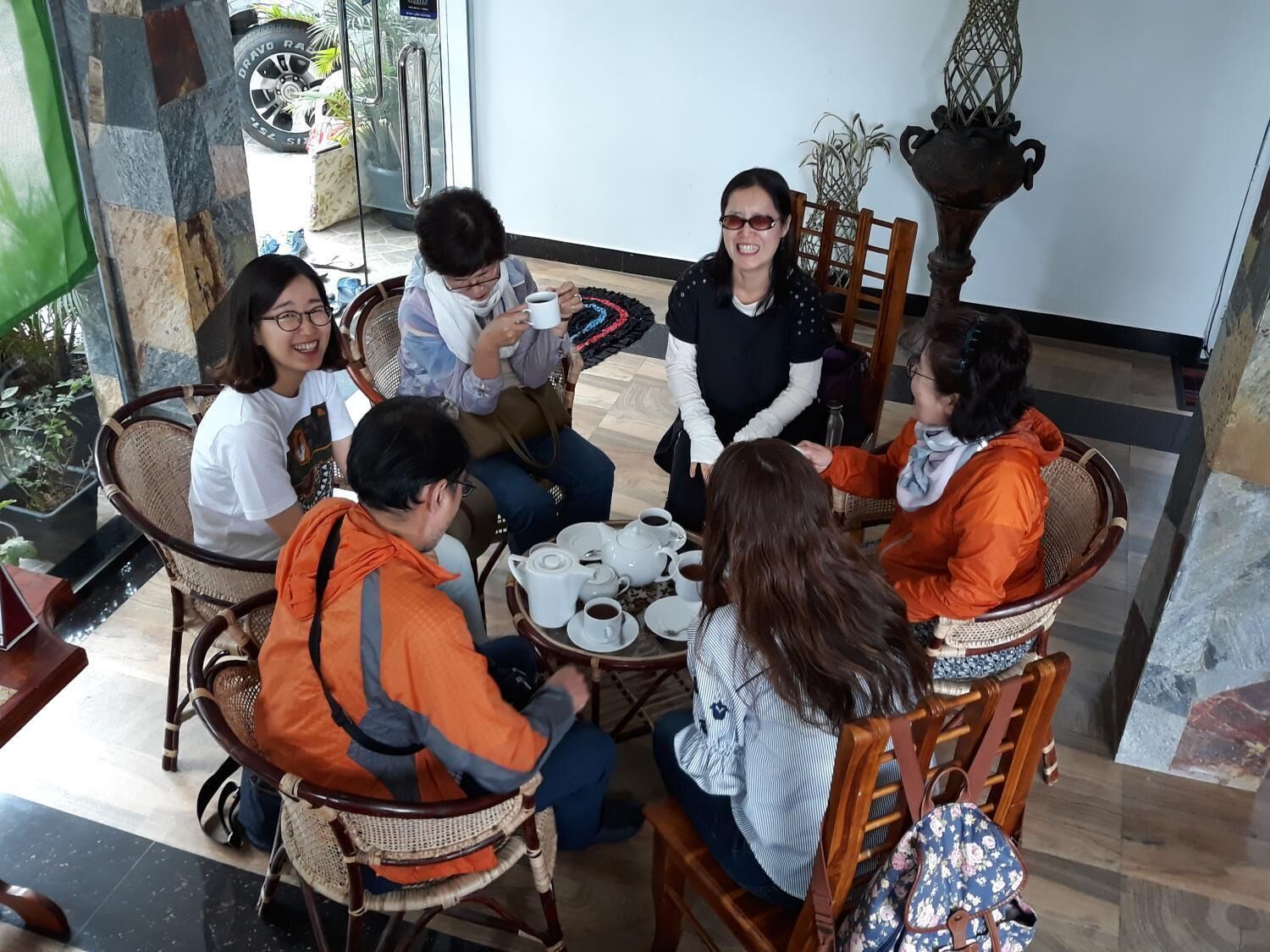 Un groupe de personnes est assis autour d’une table, devant des services à thé, dans une pièce où se trouvent des chaises en osier et un grand vase décoratif.
